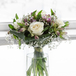 In this image, a tender sympathy bouquet rests in a clear cylindrical glass vase, as if ready to be placed on a console table in a calm Stanmore hallway after a respectful delivery. At the core of the design are several sumptuous white roses, fully open and luxuriously petalled, their creamy centres spiralling outward in soft, rounded whorls that speak of purity, love, and farewell. Around them, airy clusters of white gypsophila form a delicate veil, filling the spaces with countless tiny blooms that together resemble a fine, misty cloud, softening the overall silhouette. Interlaced among the roses and gypsophila are elegant alstroemeria stems, some still held in slender green buds, others unfolding to reveal elongated petals tinged with muted lavender and pale purple, marked by fine stripes that add subtle interest without interrupting the calm mood. Broad, dark green leaves are wrapped snugly around the lower part of the bouquet, their glossy surfaces forming a structured collar that supports and frames the flowers. Finer, bright green foliage rises through the arrangement, giving vertical movement and a gentle reminder of life continuing. Through the perfectly clear glass vase, the straight, clean stems can be seen, aligned neatly and bound just above the water with a discreet green tie. The water appears fresh and transparent, catching flecks of light from the softly glowing background. That pale, out-of-focus backdrop gives the impression of open space and quiet, much like the tranquillity found in the residential lanes and gardens around Stanmore. Overall, the bouquet offers a message of comfort and calm respect, a dignified floral expression for funerals, memorials, or condolence gestures within the local community.