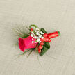 This carefully composed boutonniere showcases a single fuchsia rose, its petals velvety and richly saturated, opening in a gentle spiral that draws the eye inward like a promise whispered before a Stanmore ceremony. The rose's vivid hue is softened and framed by clusters of delicate white baby's breath, each tiny bloom adding texture and a sense of airy lightness, as if echoing the softness of a bridal veil. Around the central rose, bright red hypericum berries cluster in small groups, their rounded forms and polished sheen adding a joyful, contemporary note that contrasts with the more traditional elements, perfect for a wedding or formal event near Stanmore Broadway or a reception at a local country house. Encircling the flowers are loops of deep green foliage, carefully curved to cradle the design and create a sense of fluid movement, while slender, arching strands of ornamental grass lift the eye and contribute a fresh, natural feel reminiscent of nearby green spaces. The stems are gathered and wrapped in a matching red ribbon, wound snugly and smoothly to form a slim, elegant handle that is both practical and refined, clearly the work of an experienced florist. Positioned on a soft, neutral fabric backdrop, the boutonniere's colours appear especially striking: the bold cerise rose, pure white gypsophila, rich green leaves, and ruby-toned berries all working together in harmonious proportion. In imagining it up close, you might sense the faint, clean scent of rose petals and greenery, and feel the cool satin ribbon and firm stems beneath your fingers. It is a compact, expressive piece designed to be pinned to a lapel and worn through an important Stanmore celebration, quietly carrying sentiment and care.