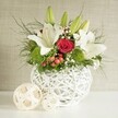 A stylish wedding-themed floral centerpiece stands on a smooth white surface, the entire arrangement framed by a softly textured beige wall that keeps attention on the flowers themselves. The focal vessel is a round, white, open-weave vase, its intersecting strands reminiscent of woven wicker or delicate twigs shaped into a modern sculpture. From this distinctive base rises a lush bouquet, dominated by large white lilies whose elegant petals open outwards, revealing gentle speckles and warm yellow stamens at their centres. Some lilies are just beginning to unfurl, their petals still hugging the buds, suggesting the slow, graceful unfolding of a wedding day in Stanmore, from quiet preparation to joyful celebration. Interspersed among the lilies are velvety red roses; one commanding rose near the centre is especially striking, its petals tightly coiled into a precise spiral, symbolising romance and devotion. Small, spherical bright green flowers-likely green dianthus or pom-pom chrysanthemums-nestle between the larger blooms, adding a vivid, lively contrast and a soft, cushiony texture that invites the imagination to reach out and touch. The arrangement is threaded with delicate stems of white baby's breath, which scatter tiny blossoms like stars across the composition, while clusters of small red berries introduce a subtle festive atmosphere, perfect for winter weddings or seasonal celebrations. A generous mix of foliage completes the design: fine, hair-like fronds extend gracefully beyond the vase, while broader, darker leaves provide structure and depth, ensuring the vibrant flowers stand out. To the left of the main vase, two decorative woven spheres in a pale natural tone rest on the tabletop, mirroring the vase's pattern and creating a sense of continuity. The overall impression is one of polished yet approachable elegance, as if crafted by Flowers Stanmore for a couple seeking a centerpiece that reflects both classic beauty and the relaxed charm of their local area.