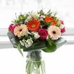 A vibrant array of fresh flowers, arranged with skill and care in a tall, clear glass vase, celebrates the colourful spirit of Stanmore. Central to the arrangement are peach and orange gerbera daisies, their velvety petals and deep brown eyes surrounded by the luxuriant spirals of magenta roses-each bloom glowing softly under gentle, natural light that might remind one of mornings near Church Road. Delicate sprays of white alstroemeria and petite roses peek out between the larger flowers, their crisp petals bringing lightness and a touch of refined elegance. Hints of dusky pink and the daintiest white blossoms contribute extra texture and depth. Framing the bouquet, lush, broad aspidistra leaves are expertly curved, cradling the arrangement and lending both protection and graceful structure, echoing the parklands that give Stanmore its unique character. Slender shoots and additional greenery intertwine between stems, which are clearly visible through the pristine glass, signifying just-picked freshness. This bouquet, infused with feelings of happiness, gratitude, and thoughtful connection, is perfect for marking a special celebration or delivering a touch of floral joy to homes across Stanmore.