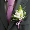 This image offers a close, intimate view of a single lapel boutonniere, pinned to the left side of a dark charcoal grey suit that has clearly been pressed and prepared with care. You can see the crisp line of the lapel, the subtle weave of the fabric, and the refined stitching that give the suit a classic, tailored feel, the kind many of our clients in Stanmore choose for weddings at local venues and nearby synagogues. Beneath the jacket lies a light dress shirt patterned with the finest of dark checks, almost a whisper of design, providing a gentle backdrop to a smooth lavender necktie. The tie hangs straight and sure, its satin surface reflecting a soft glow, conveying a sense of calm confidence. Nestled against this formal canvas, the boutonniere becomes the star. Several white alstroemeria blossoms unfurl, each petal decorated with fine reddish-brown speckling and yellow-toned centres, lending the flowers a painterly, almost watercolour quality. At the centre, a tall, conical green bud-likely tuberose-rises like a slender spire, its fresh lime tone adding an uplifting spark and a hint of future fragrance. Around it, clusters of tiny white gypsophila resemble scattered stars, intertwined with a curated mix of greenery: thin, spiky blades, softer, feather-like greens, and sturdier, broad leaves that ground the design. At the base, a narrow lilac satin ribbon wraps the stems and forms a small, precise bow, picking up the purple of the tie and completing the visual harmony. The whole composition feels cool, composed, and thoughtfully designed, the kind of boutonniere you might wear walking from Stanmore Station to a ceremony filled with anticipation and quiet joy.