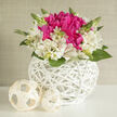 A compact yet luxurious floral arrangement in a white wicker basket appears as though it has been styled for a refined Stanmore wedding table. The design is low and rounded, its silhouette smooth and perfectly proportioned. At its heart, lustrous fuchsia blooms cluster closely together, their densely packed petals curving inwards and outwards to form ruffled, almost cushion-like shapes. Their vivid, jewel-toned colour provides a striking contrast to the ring of ivory alstroemeria surrounding them. The alstroemeria blossoms are soft and light, with slim trumpet-shaped petals that flare open gently, marked with fine touches of pink and subtle speckling that add delicate character to each flower. Interspersed among the blossoms, small buds and fresh green leaves emerge, punctuating the arrangement with hints of growth and the promise of new chapters, much like couples beginning married life in Stanmore's leafy streets.
The flowers are nestled in a round basket crafted from bright white wicker, its interwoven strands forming an intricate lattice with a modern, sculptural feel. The weave is open enough to let light pass through, casting tiny shadows on the surface beneath and emphasizing the hand-crafted nature of the piece. Two small, cream-coloured orbs made from similar woven material lie close by, echoing the texture of the basket and giving the overall composition a gentle, curated look. The lighting is soft and even, reminiscent of natural daylight streaming through large windows in a Stanmore home. The mood is polished yet warm, ideal for wedding centrepieces at local venues, civil ceremonies near Stanmore station, or elegant thank-you flowers delivered to family after the celebration. The imagined scent is fresh and green with a delicate floral sweetness, never overpowering, simply enhancing the sense of quiet joy.