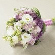 Laid elegantly upon a pale, textured surface, this sumptuous Stanmore bridal bouquet glows in an enchanting blend of soft white, lavender, and lush fresh green. Plump white Lisianthus flowers, petals gently layered and creamy, form the bouquet's centrepiece. Their graceful abundance is accentuated by sprays of unopened buds, interwoven with pastel pink and lavender Stock that ripple with delicate ruffles. Striking deep purple Allium 'Star of Persia' punctuate the arrangement with bold, spiky spheres, providing sculptural depth and contrast, reminiscent of the creative character found in Stanmore's local floristry. Slender green grasses and lively Lisianthus buds complete the design, infusing it with vitality. The bouquet's stems are bound tightly in a vivid fuchsia satin ribbon, finished with a string of soft white pearls, echoing the timeless elegance of ceremonies at St John's Church or garden parties along Stanmore's leafy avenues. Soft, even lighting reveals each bloom's intricate detail, inviting the viewer to imagine the tender scent and velvety touch of petals. An embodiment of classic taste and contemporary flair, this arrangement is ideal for weddings, anniversaries, or as the crowning centrepiece for any special event in Stanmore.