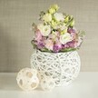 Set against a muted beige wall in a tranquil Stanmore home, this handcrafted floral arrangement draws the eye and soothes the soul with its serene palette and tactile artistry. The heart of the display is a rounded, open-weave vase of white-painted rattan branches-its airiness and intricate pattern offering a modern touch, much like the sophisticated decor found along Stanmore's Green Lane. Overflowing from this vessel is a cascade of creamy white and pale yellow eustoma, each bloom sporting gentle, ruffled layers that capture the essence of fresh spring mornings. Pink stock flowers rise gracefully amidst the eustoma, their vertical form and breath of delicate perfume infusing the bouquet with refined energy. Nestled at the base, velvety purple filler-reminiscent of local wild asters-creates lush textural depth, complemented by a rim of deep, glossy green leaves that tie the whole composition together. On the left, two artisanal spheres woven from light rattan rest on the polished table, their open, lacy structures reflecting the careful craftsmanship evident in every detail of Stanmore's local floristry. The scene is illuminated by soft natural light, which lifts each flower's pastel hue and brings warmth to the entire setting. Evocative of wedding breakfasts or sophisticated home gatherings in the Stanmore area, this bouquet is a testament to the town's gentle elegance and timeless floral style.