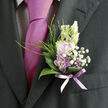 Nestled against the rich, dark fabric of a formal suit, this boutonnière radiates understated charm and the kind of quiet detail guests notice up close. At its heart, pale lavender blossoms open in small, layered rosettes, the petals thin and softly ruffled, suggesting the gentle touch of early morning light. Around them, frothy white filler flowers cluster like a breath of cloud, their tiny, star-shaped blooms adding both texture and a sense of airy romance. A ring of glossy green leaves cradles the whole piece, their curved shapes and polished surfaces providing a natural frame that keeps the florals feeling crisp and composed. Fine, grass-like strands and delicate sprigs of greenery rise and arch above the central flowers, hinting at the untamed edges of a spring garden just beyond Stanmore Hill. The stems are gathered and wrapped in a sheer lavender ribbon, tied snugly so that only a slim, elegant line of satin shows, mirroring the nearby tie and shirt collar. The colours-soft lilac, pure white, deep green, and a whisper of pastel ribbon-blend into a palette that feels perfectly suited to a wedding at a Stanmore church or a civil ceremony near Canons Park. You can almost sense a light floral fragrance following the wearer as they greet guests, shake hands, and pose for photographs along leafy local streets. Every element speaks of careful selection and expert handiwork, capturing grace, romance, and the artistry required to create something so small, yet so meaningful, for a memorable celebration.