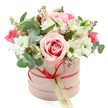 A refined floral hatbox arrangement is presented against a crisp white background, the soft colours and balanced composition reflecting the gentle, residential charm of Stanmore. The box itself is a round, cylindrical shape in a muted pastel pink, with a smooth matte finish that looks velvety to the touch. A thin coral-red satin ribbon wraps around the middle and ties at the front in a small, carefully formed bow, from which a cream gift tag with green script and a discrete website address hangs, suggesting this is a planned, delivered gift rather than a last-minute purchase. Above the rim of the hatbox, the bouquet forms a lush, domed canopy of flowers in shades of pink and white, enriched by cool greens. A standout rose sits prominently at the front, its fully opened petals displaying a gentle bi-colour effect: pale pink on the outer edges, deepening to a warm rosy swirl at the centre. Around it, white daisy-like chrysanthemums with fresh green centres brighten the arrangement and bring a sense of easy cheer. Threaded throughout are stems of white alstroemeria, their trumpet-shaped blooms accented with fine speckled markings, while pockets of smaller intense pink flowers-likely spray roses or lisianthus-add depth and a hint of drama. The greenery is thoughtfully interwoven, with rounded, silver-green eucalyptus leaves providing a contemporary, airy feel, supported by darker green foliage and delicate stems tipped with small green buds. The lighting is clear and soft, enhancing the natural colours and the sense of freshness. This bouquet feels tailored for special occasions-birthdays, anniversaries, thank-you gifts-delivered to homes near Stanmore Hill, quiet streets off Uxbridge Road, or family gatherings around The Broadway, carrying a message of warmth and considered elegance.