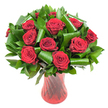 A close, sensory portrait of a lush, rounded bouquet built around a dozen or more deep crimson roses, each bloom open to reveal velvety, spiralled petals that catch the light like fine silk. The roses' colour leans toward a wine-dark red, saturated and warm, their edges soft and unblemished as if just cut from morning stems. They sit nestling in a generous halo of glossy, dark green foliage-dense, flat leaves that read as Salal or Italian Ruscus-whose smooth, reflective surfaces create a cool contrast to the tactile velvet of the petals. Interspersed among the greenery are broader, emerald leaves carefully rolled and folded into sculptural coils and loops, their architectural curves adding a contemporary rhythm to the classic rose silhouette. The whole composition rises from a tall, translucent ruby-red glass vase with a subtle ribbed or twisted texture that throws faint vertical highlights and deepens the overall red palette; the vase's slender waist and flared mouth balance the bouquet's volume with elegant poise. Soft, even studio lighting isolates the arrangement against a pristine white background, making the reds and greens read with amplified clarity and giving the scene a calm, luminous stillness. Close your eyes and imagine the faint, heady fragrance of fresh roses and the weight of careful florist wiring-this is an arrangement that speaks of considered craft, classic romance, and quiet luxury. Perfect for an intimate anniversary, a refined Valentine's gift, or a centerpiece that anchors a dining table with passionate, understated style in and around Stanmore and nearby Canons Park.