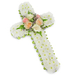 A beautifully crafted white floral cross is presented against a bright, uncluttered background, its gentle shadow giving it a quiet sense of presence. The entire form of the cross is richly covered in small white button chrysanthemums, each bloom pressed closely against its neighbours so that the surface appears almost like a padded quilt of petals. This dense, snowy covering radiates purity and stillness, creating a soothing focus for the eye. Running carefully around the outer edge is a narrow band of lime green ribbon, wrapped in neat, even turns that follow the contours of the cross, providing a subtle frame and a fresh flicker of colour. At the very centre, where the upright and crossbar meet, lies a delicate cluster of roses and foliage. The roses are fully open and softly rounded, in shades of blush pink, warm peach, and creamy ivory, their colours blending in a gentle, harmonious palette. Light green fern fronds emerge from beneath the blooms, while tiny lavender-hued filler flowers peek through here and there, adding fine texture and a quiet hint of contrast. The lighting is soft yet clear, ensuring that the details of each petal and ribbon fold are visible while maintaining a serene atmosphere. This floral cross feels like a tender, respectful tribute, the kind a Stanmore family might have arranged for a church altar, crematorium service, or graveside farewell, offering a visible expression of sympathy, faith, and cherished remembrance during a time of mourning.