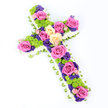 A lush, cross-shaped funeral flower arrangement is presented on a stark white background, its form gently angled so that the top seems to lift slightly toward the viewer while the base recedes. The outer edge of the cross is meticulously framed by a thick ring of white spray chrysanthemums, creating a smooth, cushion-like border that suggests purity, peace, and comfort. Just visible behind this white edge is a fine band of folded light green ribbon or foliage that wraps around the entire perimeter, adding a delicate whisper of green and showcasing a florist's careful finishing touch. Inside the border, the cross is filled with an intricate mix of blooms. Large roses in shades of bold fuchsia pink and creamy pale yellow dominate the design, their generous, open petals conveying both beauty and tenderness. Between these roses, bright lime-green button chrysanthemums stand out, their compact, spherical heads contributing lively texture and a fresh, contemporary note. Smaller flowers in deep purple and violet tones-likely lisianthus or statice-thread through the arrangement, filling gaps and adding depth, while also lending a more reflective, spiritual mood. Soft, even lighting highlights each flower without casting harsh shadows, ensuring the colours remain vivid yet suitable for a solemn occasion. Crafted with the thoughtful attention typical of Flowers Stanmore, this floral cross would be a fitting tribute for a local funeral or memorial service, whether placed beside the altar in a Stanmore church or resting gently at a graveside, symbolising faith, remembrance, and the enduring love shared with the person who has passed.