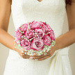 The photograph focuses closely on a bridal bouquet held just in front of an intricately laced ivory wedding dress, evoking the calm elegance of a Stanmore ceremony. The bouquet is tightly gathered into a rounded, hand-tied design, made up of fully open blush-pink roses, each one perfect in form. Their petals unfurl in soft, concentric layers, giving the bouquet a plush, almost cushion-like appearance. Nestled between the roses are light, airy clusters of white gypsophila, their hundreds of tiny flowers creating a misty veil that softens the outline of the bouquet. The contrast of pink and white is gentle rather than stark, blending into a harmonious palette that feels ideal for a classic wedding at a local Stanmore venue, perhaps near Stanmore Hill or along The Broadway. The bride's hands are relaxed yet purposeful, fingertips resting lightly on the stems, suggesting a sense of serenity and readiness as she prepares to walk down the aisle. The lighting is soft and flattering, reminiscent of natural daylight streaming through church windows, casting a warm glow over the blooms and catching the subtle textures of both the flowers and the lace. You can almost sense the quiet rustle of the gown and the faint, fresh scent of roses, like stepping into a beautifully prepared reception room at a nearby country club. This bouquet's balanced composition and delicate colour story evoke enduring romance, making it feel not just like an accessory, but like a floral expression of the bride's emotions on her wedding day in Stanmore-hopeful, reflective, and lovingly composed.
