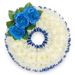 A bespoke circular sympathy wreath crafted in the classic shape of the numeral '0', photographed from directly above on a clean white background. The foundation is a dense, cushion-like sea of small white spray chrysanthemums - hundreds of tiny florets tightly packed to form a smooth, tactile surface that reads as calm and consoling. Along the upper-left curve a cluster of three royal blue roses anchors the design; their velvety petals unfurl from firm centers, the deep sapphire tone almost electric against the pale chrysanthemum field. Glossy dark green foliage fans discreetly around the roses, adding natural depth and a fresh, slightly herbaceous scent beneath the sweeter rose perfume. Both the inner and outer rims of the figure are traced with a pleated royal blue satin ribbon, ruffled to produce a scalloped silhouette that repeats the roses' vivid hue and gives the tribute a formal, finished edge. Soft studio light throws gentle shadows into the flower textures, accentuating the chrysanthemum's fluff and the satin's sheen without harsh contrast, so the overall feeling is one of dignified stillness and care. This personalised numeric wreath is often ordered as part of a numbered tribute for memorials or funerals to mark an age or special date, offering a quiet, tactile way to honour memory. As a local Stanmore florist might explain, the cool blue and pure white read as peaceful and respectful-thoughtful colours for a farewell in a neighbourhood near Stanmore High Street or Canons Park.