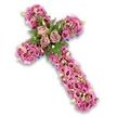 In this image, a lavish floral cross is carefully centred on a bright white background, making every tone of pink, mauve, and purple stand out with clarity. The cross is fully three-dimensional, yet no foam or base can be seen; instead, a continuous layer of blossoms covers the entire surface. At the point where the arms meet, three large roses in soft dusty rose and light mauve shades create a calm, inviting centrepiece. Their open faces and velvety petals lend the design a sense of gentleness and grace. Embracing these roses, fronds of fern and small, neat green leaves provide a cool, refreshing counterpoint to the warm pinks. Moving outward from the centre, the arms and base of the cross are filled with an abundance of lisianthus blooms, their ruffled petals and trumpet shapes giving the arrangement movement and texture. The colours shift subtly from pale blush to more vivid pinks with darker centres, like soft ripples across the surface. Interspersed among the lisianthus are tiny sprays of pink and purple accent flowers, touches of pale green buds, and a few white blossoms that glimmer softly against the more saturated tones. Along the very edge of the cross, narrow hints of dark purple ribbon or fabric peep out, gently framing the silhouette and adding depth. The overall effect is one of serene beauty and solemn respect, a tribute that feels both personal and timeless. It is the kind of floral cross a family in Stanmore might choose to send to a funeral or memorial, trusting that the flowers speak of love, faith, and remembrance when words feel too small.