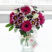 A vibrant mixed bouquet of fresh flowers stands proudly in a low, textured clear glass vase, placed against a bright, softly blurred white backdrop that feels like daylight pouring through a Stanmore bay window. The arrangement is dominated by large gerbera daisies in deep fuchsia and magenta, their velvety petals circling dark, almost chocolate centres that draw the eye immediately. Around them, bicolour chrysanthemums unfold in layers of rich purple at the petal tips, gradually fading to white towards the centre, where small yellow discs glow like tiny suns and add a playful sparkle. Creamy white roses, some fully open and others still in tight buds, nestle between the bolder tones, adding a gentle, romantic balance and hinting at a soft, sweet fragrance. Fine sprigs of white gypsophila, or baby's breath, float throughout the bouquet like delicate lace, creating a light, airy texture that softens the strong colours. Deep green foliage fans out from the base and peeks between the blooms, giving the arrangement structure and a fresh, garden-picked feel, as if the flowers had just been gathered from a well-tended plot near Stanmore Country Park. The vase itself is tied around the neck with a simple rustic twine bow, the natural fibres slightly frayed, suggesting care and a handmade touch. A small, pale rectangular gift tag hangs from the twine, decorated with a soft pink floral motif and subtle lettering that hints at a personalised message, even if the words are not legible. Bathed in gentle, natural light, every petal and leaf appears crisp and dewy, evoking the joy of receiving a surprise flower delivery in Stanmore to celebrate a birthday, anniversary, or just to brighten a quiet afternoon at home.