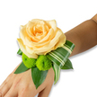 A delicate wrist corsage rests gently on the inner forearm of a person, photographed against a bright, plain white background so every petal and leaf reads clearly. At the heart of the piece sits a single, fully opened peach-apricot rose, its concentric petals soft and satiny, folding inward in a warm spiral that catches a subtle, diffused light. Just below and slightly to the left are two lime-green button chrysanthemums, their compact, pom-pom heads offering a lively, textured counterpoint to the rose's smooth curves. The flowers are cradled on a bed of deep green foliage - broad, glossy leaves, likely salal or ruscus, that create a dense, supportive base and give the wristlet a grounded, natural profile. A variegated, striped leaf has been artfully folded and curled from beneath the rose, its lighter and darker green bands forming a crisp, architectural line that sweeps toward the right edge of the wrist, lending a contemporary accent to the classic palette. The corsage sits close to the skin, the fastening hidden from view, so the composition appears to float like a small garden on the arm. There is a faint, clean scent of rose and cut greenery suggested by the arrangement, an intimate fragrance that suits quiet mornings before a wedding or the nervous joy of prom night. As a Stanmore florist I imagine this worn on Concorde Road or walking past Stanmore Common: soft peach and lime green catching the light while loved ones gather. The overall feeling is refined and celebratory, a carefully balanced study in texture, colour, and movement meant to accompany meaningful moments.