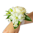 A delicate wrist corsage of soft white blooms rests gently on a fair-skinned wrist, presented against a clean white backdrop so every texture reads clearly. At its center sits a creamy ivory rose, petals velvety and slowly unfurling in a classic, romantic spiral that catches a soft, cool light and throws faint, silvery shadows. Below and offset to the left, a trumpet-shaped white bloom - suggestive of Alstroemeria or a slender lily - opens with a pale green-yellow throat that gives the composition subtle depth and a quiet, fresh contrast. Scattered through the cluster are tight green buds, poised to bloom, and tiny cloud-like sprigs of Baby's Breath that lend an airy, almost powdered texture to the silhouette. A small spray of white daisy-like florets with bright yellow centers nestles toward the lower edge, adding a cheerful, vintage touch. Broad, glossy deep-green leaves frame the arrangement, their waxy surfaces and rounded shapes amplifying the whiteness of the flowers while grounding the design with a natural heft. The corsage is secured with a multi-strand pearl bracelet of uniform white beads that wrap smoothly around the wrist, their cool lustre complementing the flowers' soft sheen and offering a tactile contrast to the blossoms. Close up you can imagine a faint, fresh scent - a clean floral whisper that suits ceremonies and formal nights alike. Designed with classic proportions and subtle modernity, this wristlet reads as refined and celebratory, perfect for weddings, prom nights, or any meaningful gathering where understated elegance is desired.