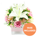 A square glass vase filled with white lilies and rosy blooms is displayed against a spotless white background, giving the entire arrangement a bright, contemporary feel. Two fully opened white Oriental lilies rise from the centre, their broad petals gently curving back as they frame the dark reddish-brown stamens that stand proud inside each flower. Clustering closely around these lilies are numerous pink roses, in tones that range from soft blush to a richer mid-pink. Some roses appear as tight, perfectly wound buds, while others are open and layered, with velvety petals that suggest both delicacy and fullness. Between the roses, slim stems of alstroemeria weave through, their petals in shades of pale pink and white, touched with yellow and fine speckles that create a sense of detailed, natural artistry. Light sprays of white baby's breath punctuate the design, offering tiny, cloud-like blooms that soften and unify the composition. Surrounding foliage in rich green tones, including broad, reflective leaves and feathery fronds, gives structure and a freshly gathered quality, as though the flowers had just been arranged by an experienced florist in Stanmore after a morning delivery. The stems rest neatly in a clear, cube-shaped vase, with the clean lines of the glass adding a modern edge. Around the midpoint of the vase, a pale pink satin ribbon is wrapped in a smooth horizontal band, providing a subtle, romantic finishing touch. In the lower right corner of the image, a bold orange circular label with crisp white capital text reads VASE INCLUDED, clearly indicating that the glass vase is part of the bouquet. The overall impression is of a ready-to-place centrepiece, perfect for celebrating milestones, expressing thanks, or simply brightening a Stanmore home with gentle elegance.